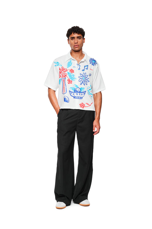 Männliches Model trägt das Crayon Summer Shirt Graphic mit skizzenhaften roten und blauen Doodles, kombiniert mit weiten schwarzen Hosen und weißen Sneakers.
