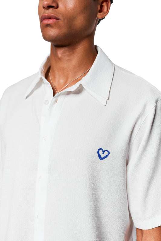 Person trägt das Crayon Summer Shirt Heart: Weiß, kurzärmelig, mit blauer Herzstickerei links. Kubanischer Kragen, dünner Stoff, entspannte Passform.