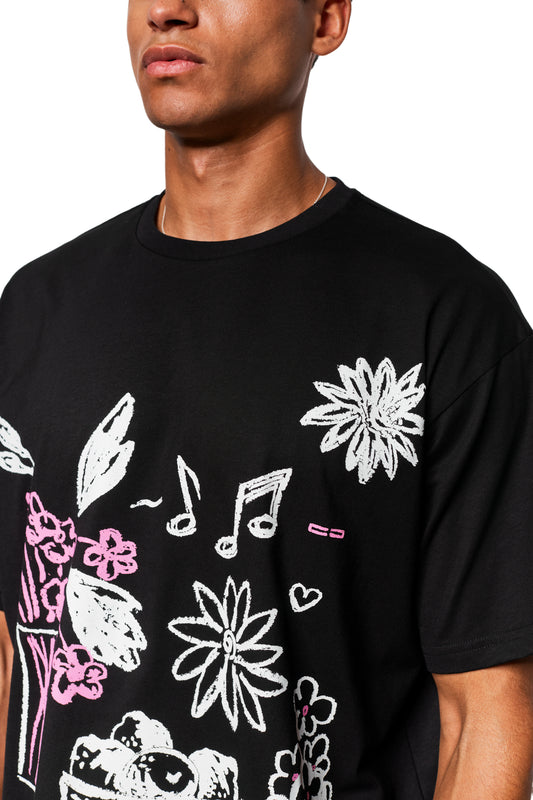 Person trägt ein Crayon T-Shirt Oversized Black mit weiß-pinken Doodle-Grafiken, inklusive Blumen und Noten, in sehr lockerer, cropped Passform.