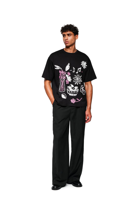 Männliches Modell trägt Crayon T-Shirt Oversized Black mit weißen und rosa Skizzen, kombiniert mit weiten schwarzen Hosen und schwarzen Schuhen, vor weißem Hintergrund.