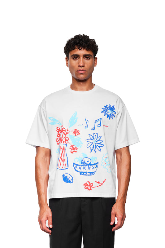 Junger Mann trägt Crayon T-Shirt Oversized White mit handgezeichneten Blumen- und Fruchtmotiven in Rot und Blau, lockere Passform, vor weißem Hintergrund.