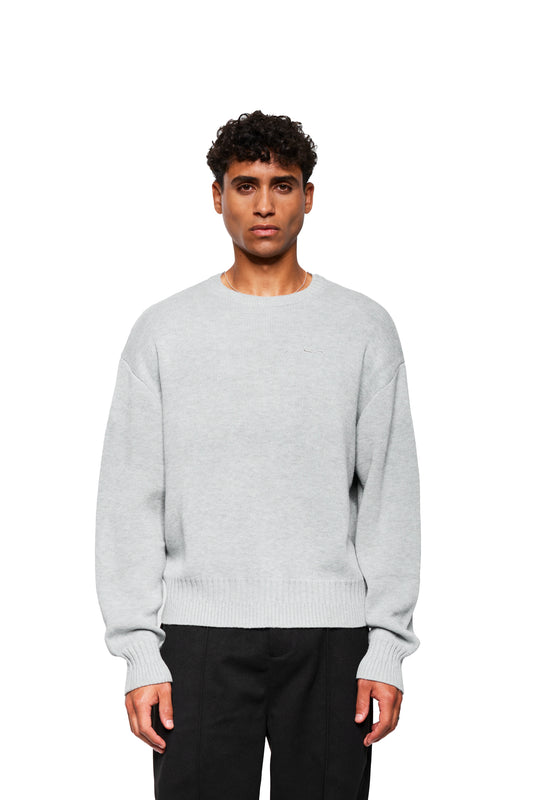 Young man wears Exquisite Knit Sweater Gray with embroidered OLAKALA icon, loose fit and different knit patterns on cuffs and collar.