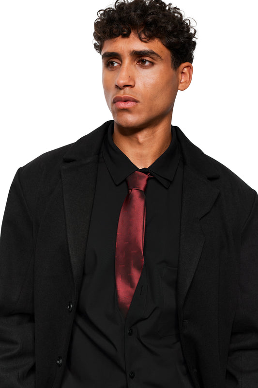 Young man in a black outfit wears a Bordeaux monogram tie by OLAKALA, with a burgundy silk sheen, against a white background.