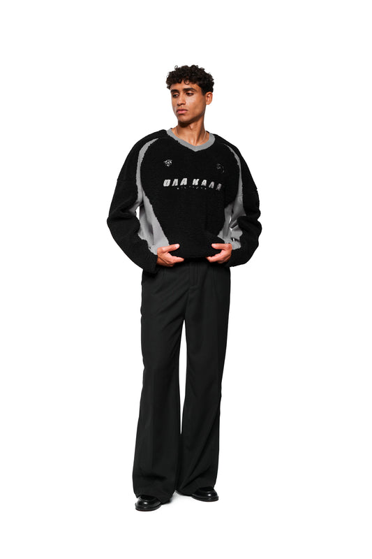 Junger Mann trägt oversized Fleece V-Neck Sweater Black mit gesticktem OLAKALA Stuttgart Logo, Isolation Jersey- und Sports-Icons, kombiniert mit weiten, schwarzen Hosen und Schuhen.