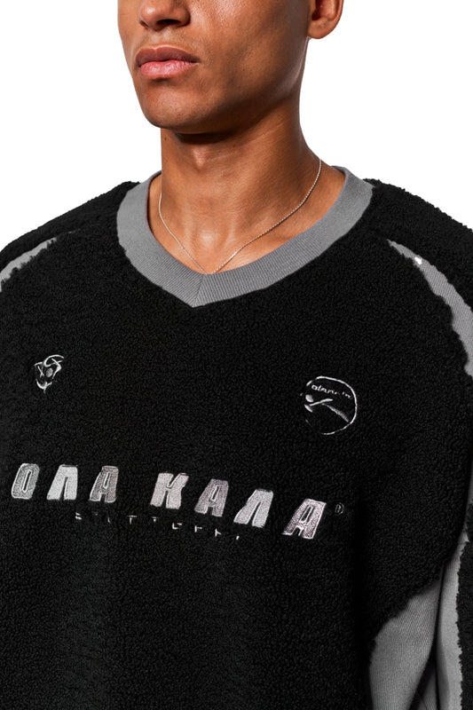 Mann trägt OLAKALA Fleece V-Neck Sweater Black mit gesticktem Logo und Icons, silberner Kette, elastischem Saum und Oversize-Passform.