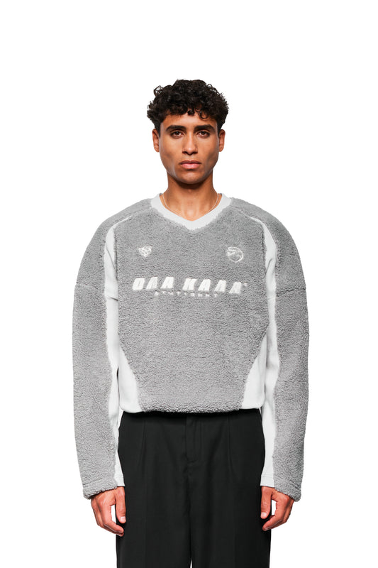 Männliches Model trägt grauen Fleece V-Neck Sweater mit gesticktem OLAKALA Stuttgart Logo, weißen Seitenpaneelen und Kordelzugsaum. Oversized, unisex Design.