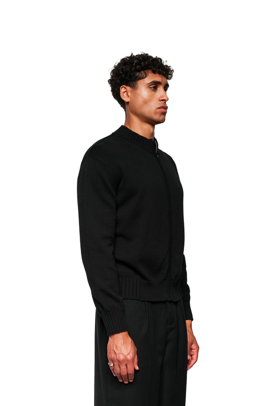 Junger Mann in schwarzer Summer Knit Jacket Black mit asymmetrischer Vorderseite und Inside-Out-Nähten, kombiniert mit lockeren schwarzen Hosen.