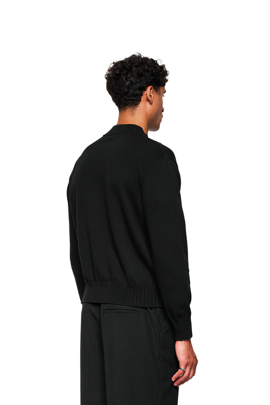 Männliches Model von hinten in schwarzer Summer Knit Jacket mit asymmetrischer Vorderseite und lockerer Passform, kombiniert mit locker sitzender schwarzer Hose.