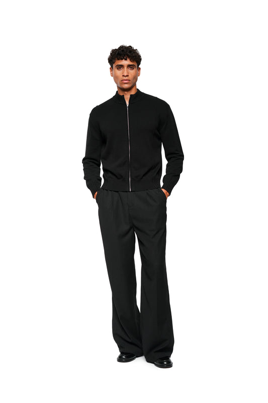 Männermodell trägt eine schwarze, asymmetrische Summer Knit Jacket mit Metallreißverschluss und strukturiertem Baumwollstrick, kombiniert mit schwarzen, weiten Hosen und Lederschuhen.