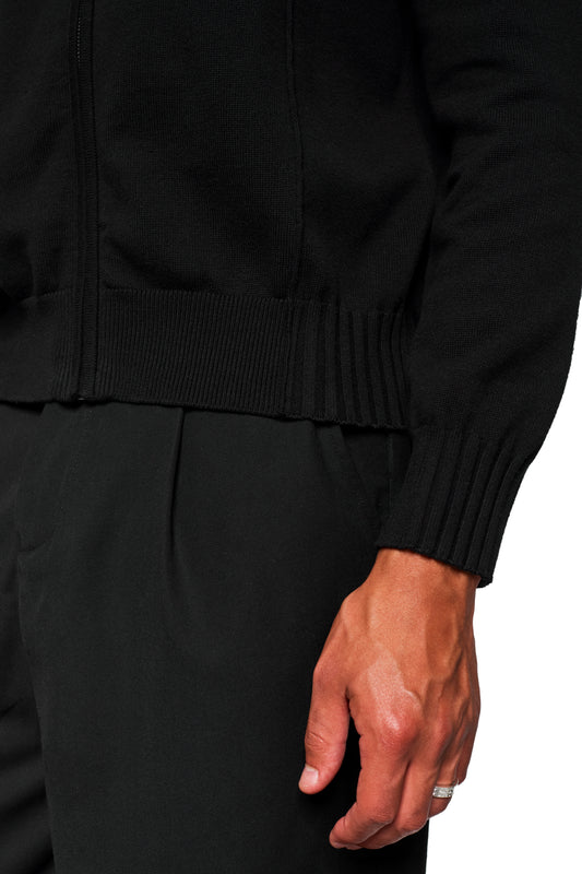 Person trägt Summer Knit Jacket Black, eine leichte Strickjacke mit asymmetrischer Vorderseite, Metallreißverschluss und strukturierter Baumwolle, passend für warme Sommerabende, von OLAKALA.