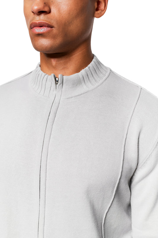 Person trägt eine hellgraue Summer Knit Jacket mit asymmetrischer Vorderseite, Rippenmuster und Metallreißverschluss, ideal für warme Abende, aus weichem Baumwollstrick.