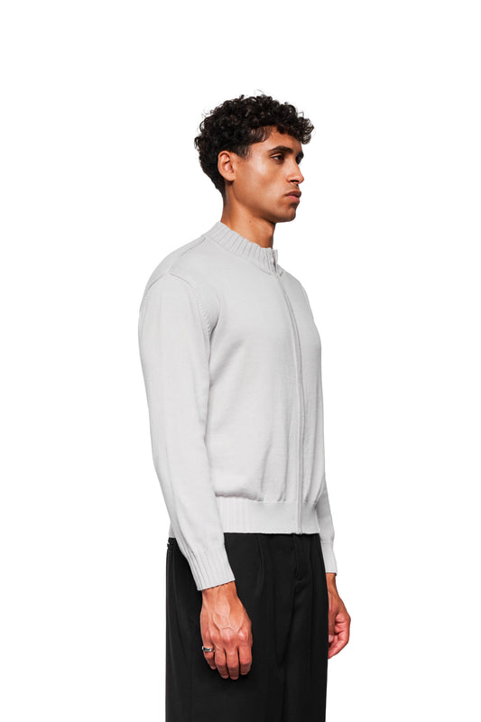 Männermodell trägt die Summer Knit Jacket Light Gray, eine leichte Strickjacke mit asymmetrischer Vorderseite und Metallreißverschluss, kombiniert mit schwarzen Hosen.