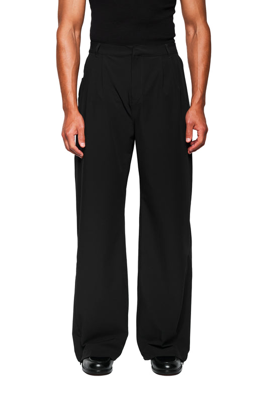 Person in loose-fitting Exquisite Tech Pants Black with asymmetric pocket openings and pleats, worn with a black sleeveless top, over polished shoes.