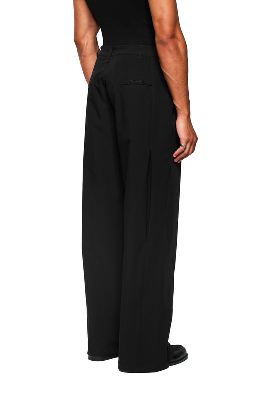 Back view of a model in the Exquisite Tech Pants Black, with asymmetrical pocket opening, pleats, embroidered OLAKALA icon and robust, stretchy material for comfort.
