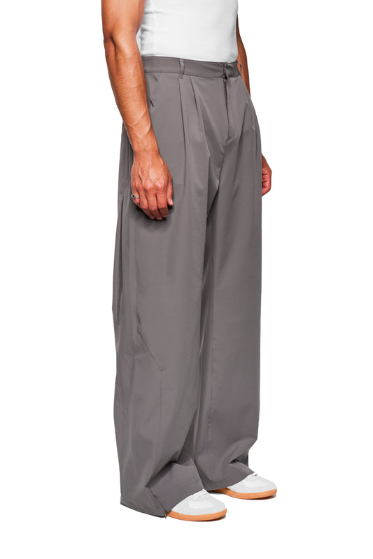 Person wears Exquisite Tech Pants Gray with pleats and asymmetrical pocket opening, combined with white sneakers. Simple silhouette emphasizes OLAKALA's unisex design.