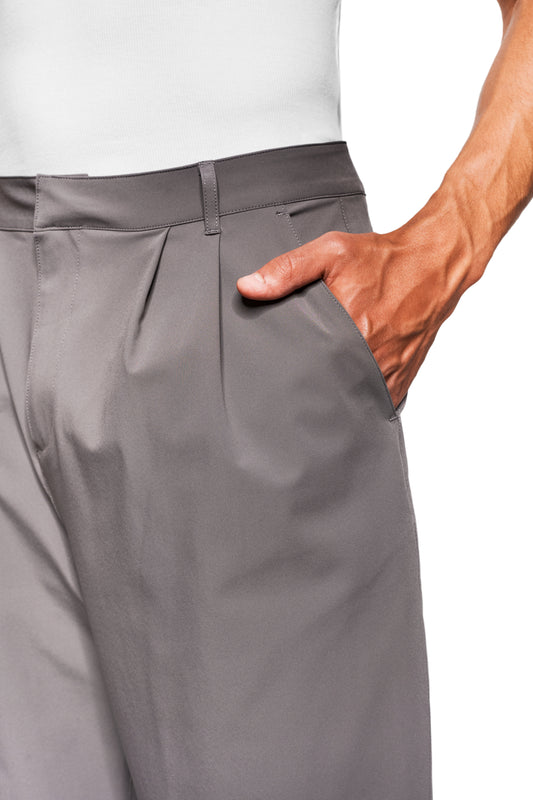 Person wearing Exquisite Tech Pants Gray with asymmetrical pocket opening and pleats, hand in pocket. OLAKALA icon visible above the right back pocket.