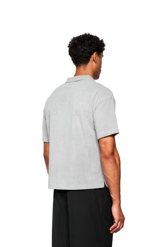 Rückansicht einer Person in einem grauen Terry-Poloshirt mit Logo-Stickerei auf der Brust, kombiniert mit locker sitzenden schwarzen Hosen.