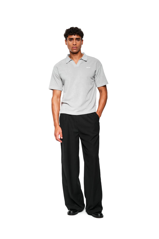 Mann trägt ein Terry Polo Gray aus weichem Frotteematerial mit Logo-Stickerei, kombiniert mit schwarzen Hosen und Schuhen, stehend vor weißem Hintergrund.