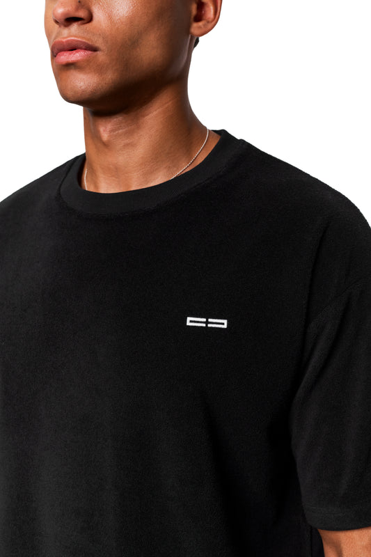 Mann trägt Terry T-Shirt Black aus weichem Frotteematerial mit weißem Logo auf der Brust und silberner Halskette. Unisex, regulärer Schnitt.