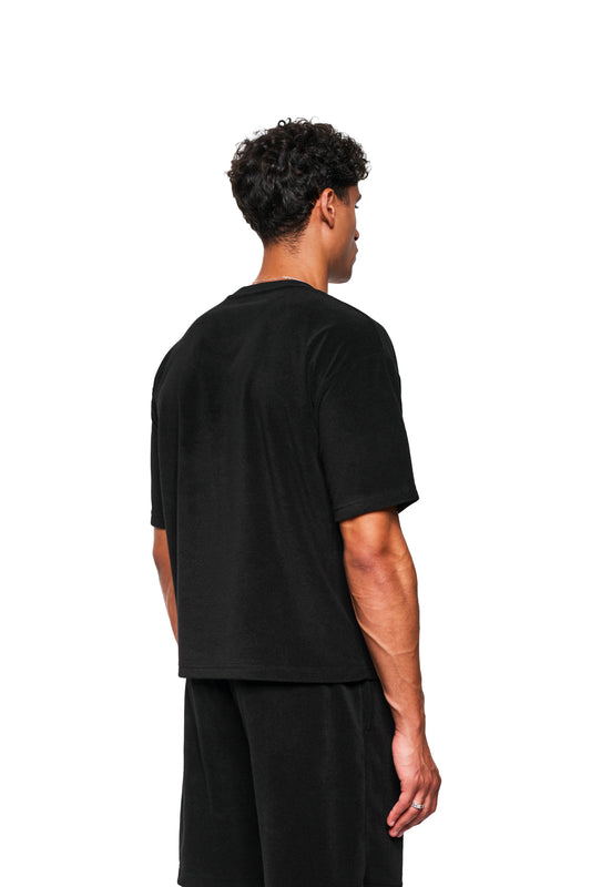 Rückansicht: Modell trägt OLAKALA Terry T-Shirt Black aus strukturiertem Frottee, regulärer Schnitt, mit Logo-Stickerei auf Brust. Kombiniert mit passenden schwarzen Shorts.