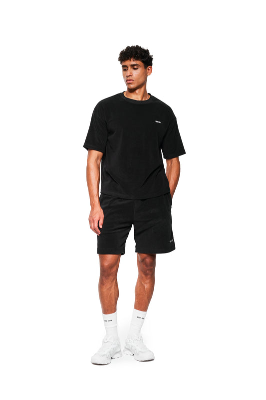 Junger Mann in schwarzem Terry T-Shirt mit Logo-Stickerei, getragen mit passenden Shorts, weißen Socken und Sneakers. Perfekt für einen lässigen Athleisure-Look.