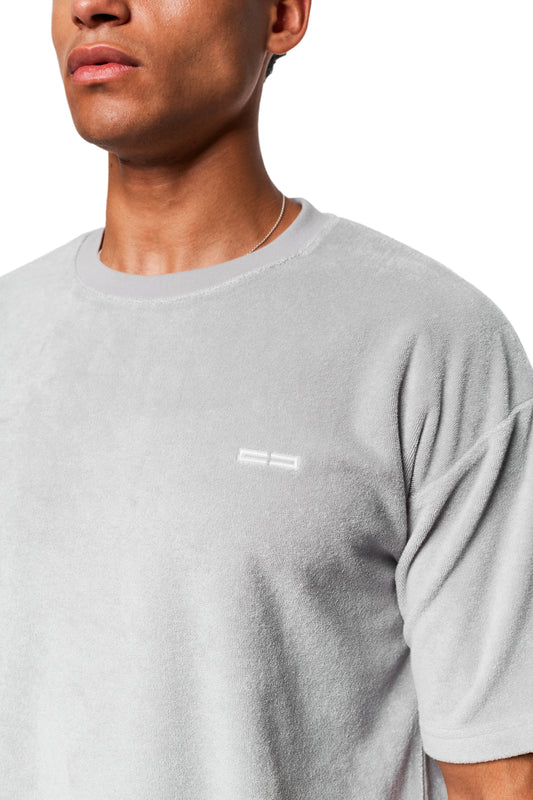 Person trägt ein Terry T-Shirt Gray aus weichem Frotteematerial mit Logo-Stickerei auf der Brust und silberner Halskette.