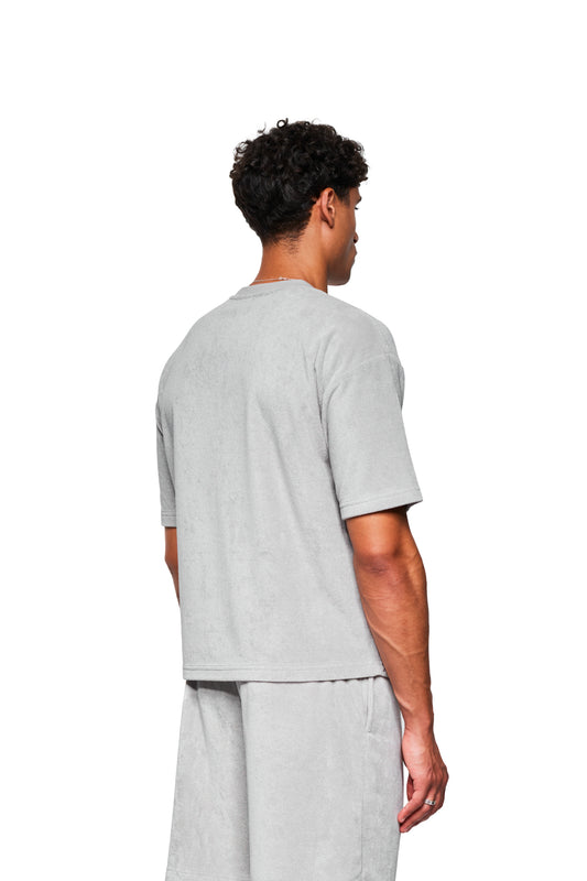 Ein junger Mann trägt ein lockeres, grau meliertes Terry T-Shirt mit Stickerei auf der Brust, passend zu Shorts, ideal für lässige Freizeitkleidung.