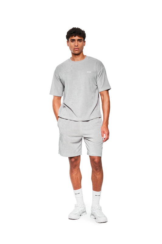 Junger Mann trägt ein Terry T-Shirt Gray aus weichem Frotteematerial mit Logo-Stickerei, kombiniert mit passenden Shorts und weißen Sneakers.