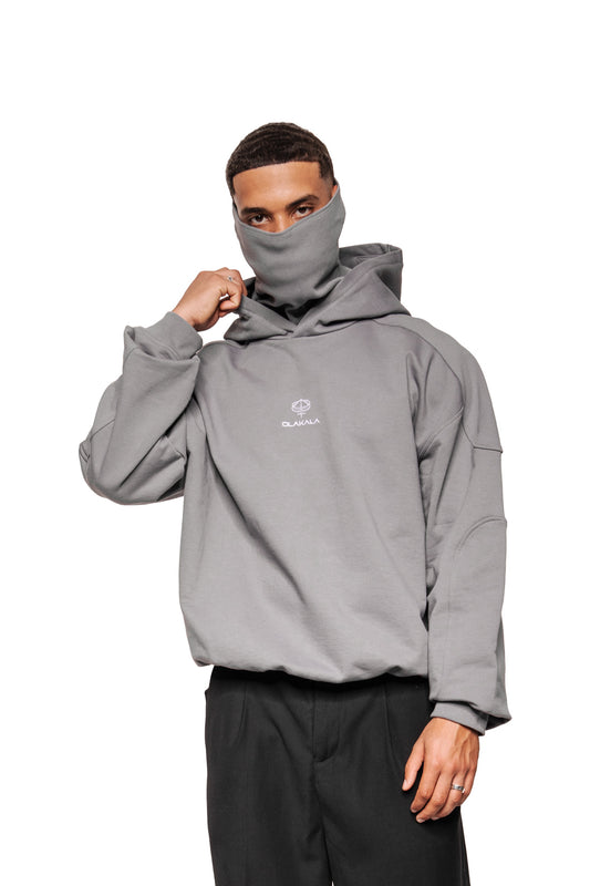 Modell trägt Discreet Mask Hoodie Dark Gray mit integriertem Hals-Gaiter und gesticktem OLAKALA-Logo, kombiniert mit weiten schwarzen Hosen.