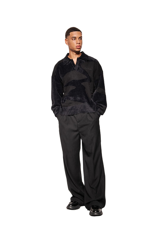 Young man in Glitched Fuzzy Polo Black with polo collar, embroidered Synergy logo and fluffy material, combined with black pants and leather shoes.