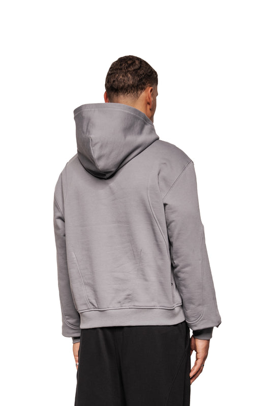 Person from behind in a Greyzone hoodie dark gray with hood at the bottom, loose-fitting black pants, accentuated by ribbed cuffs and dividing seams, against a neutral background.