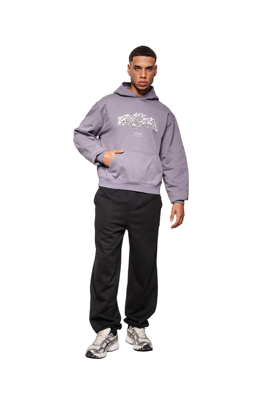 Young man wears Greyzone Hoodie Lilac with abstract graphic print, kangaroo pocket and ribbed cuffs, combined with black sweatpants and gray sneakers.