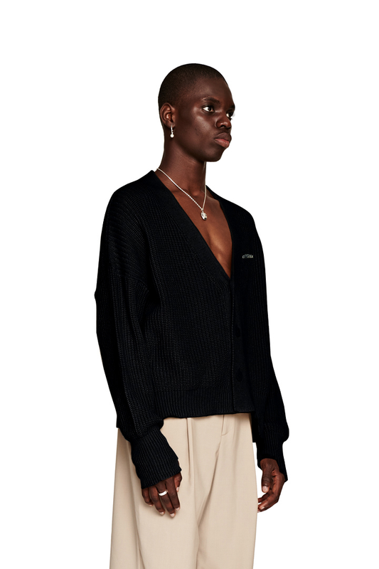 Person trägt die CC Cardigan Black mit V-Ausschnitt und versetztem Saum, kombiniert mit weißer Hose, zeigt OLAKALA Pin und oversized, cropped Design.