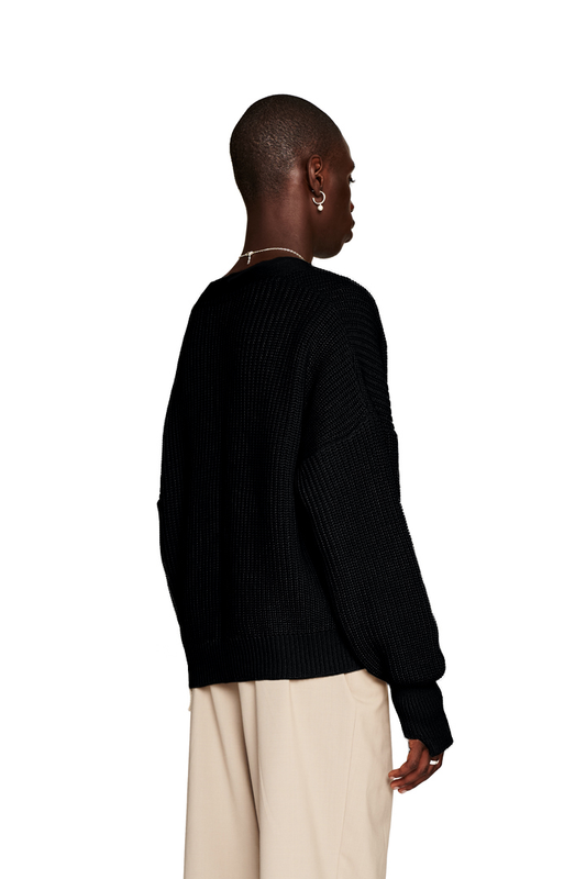 Person trägt eine CC Cardigan Black mit V-Ausschnitt, versetztem Saum, Metall-Pin und oversized, cropped Passform. Unisex-Design von OLAKALA.