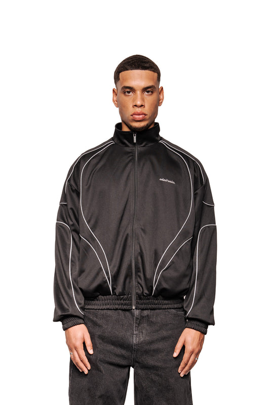 Modell trägt Rave Trackjacket Black mit Stehkragen, reflektierenden Paspelierungen, elastischen Bündchen und gesticktem OLAKALA-Logo, kombiniert mit lockeren Jeans.