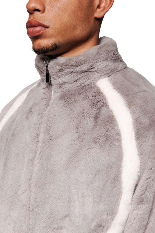 Person wears Two-Toned Fur Jacket Gray with a high collar, raglan sleeves and a stripe on the shoulder. Loose fit, with zipper and pockets.