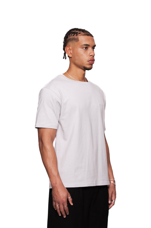 Junger Mann trägt NC T-Shirt Gray Regular mit schwarzem OLA KALA Logo auf der Brust, Rückengrafik Neighbor's Cherries, kombiniert mit schwarzen Hosen und Schmuck.
