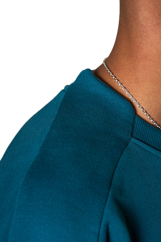 Person trägt den Defect ID Sweater Petrol mit Ripp-Bündchen und Teilungsnähten am Ärmel, Oversized-Passform und digitalem Defect Identity-Grafikdruck auf der Brust.
