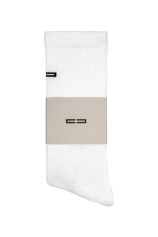 Einzelne weiße, gerippte Crew-Socke Linen Socks White mit gesticktem OLAKALA-Logo nahe dem Bündchen, gehalten von einem hellbraunen Papierband.