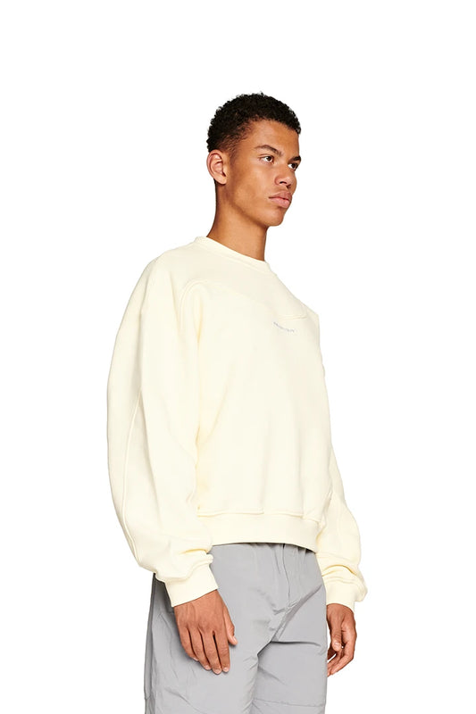 Man in the white Elysium Sweater Butter with ribbed collar, oversized fit, Elysium logo on the chest, dividing seams and ribbed cuffs.