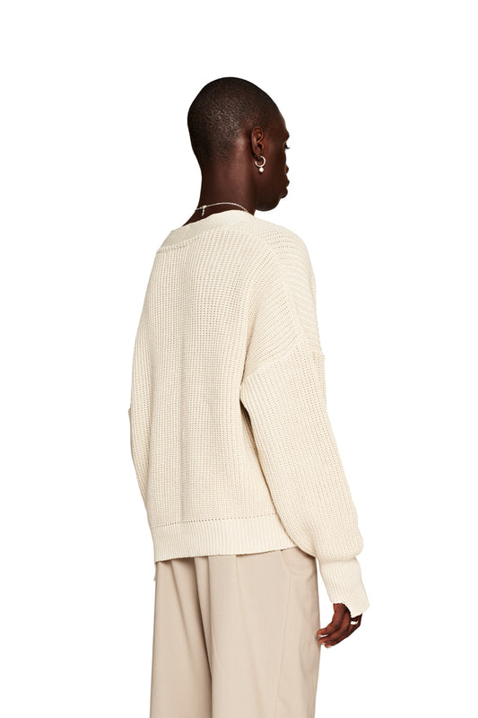 Person trägt eine weiße CC Cardigan Beige mit V-Ausschnitt und versetztem Saum, oversized und cropped, mit OLAKALA Pin, unisex Design.