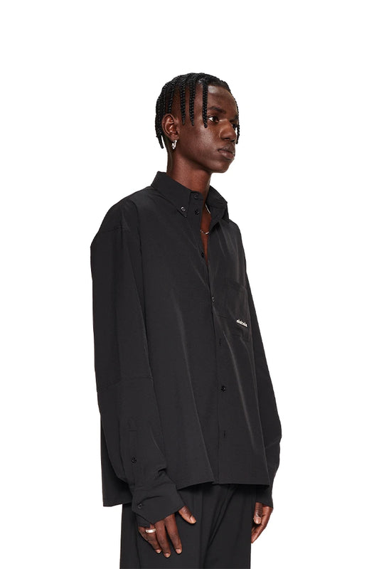 Man in the CC Button Shirt Black with double collar, chest pocket with OLAKALA metal logo and dividing seams, presents the oversized unisex fit.