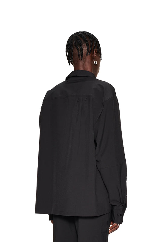 Woman wears the CC Button Shirt Black with double collar, breast pocket and OLAKALA metal logo. Oversized fit, unisex, visible dividing seams and pleats on the back.