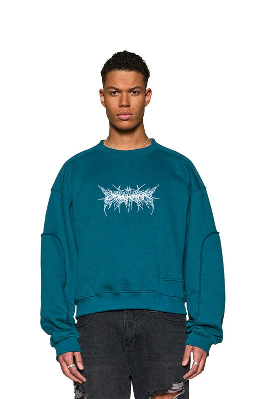 Mann trägt Defect ID Sweater Petrol von OLAKALA mit Oversized-Passform, Digitaldruck-Grafik auf der Brust und speziellen Nähten am Halsausschnitt.