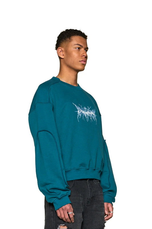 Mann trägt Defect ID Sweater Petrol mit Ripp-Bündchen und OLAKALA-Grafik auf der Brust, oversized und unisex gestaltet.