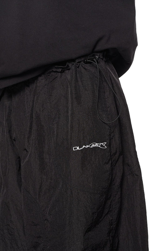Person trägt weite schwarze Parachute Pants mit seitlichen Eingriffstaschen und elastischem Kordelzug, gesticktem Synergy-Logo, aus strapazierfähigem Material.