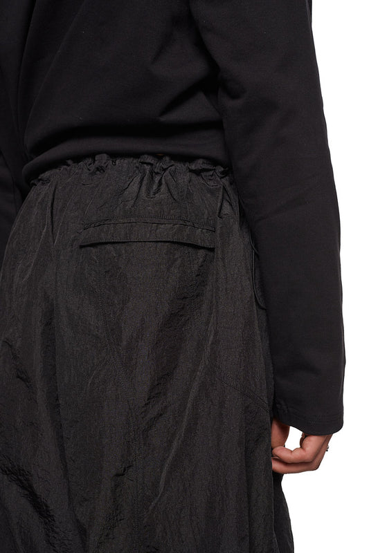 Person trägt Parachute Pants Black mit Seitentaschen, elastischem Kordelzug, gesticktem Synergy-Logo und lockerer Passform.