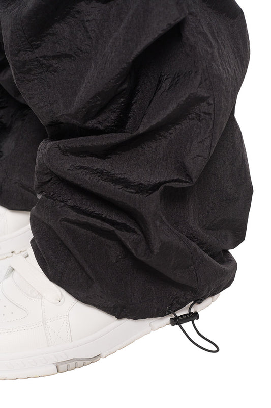 Parachute Pants Black mit elastischem Kordelzug und Synergy-Logo, kombiniert mit weißen Sneakers. Lockerer Schnitt, strapazierfähiges Material.