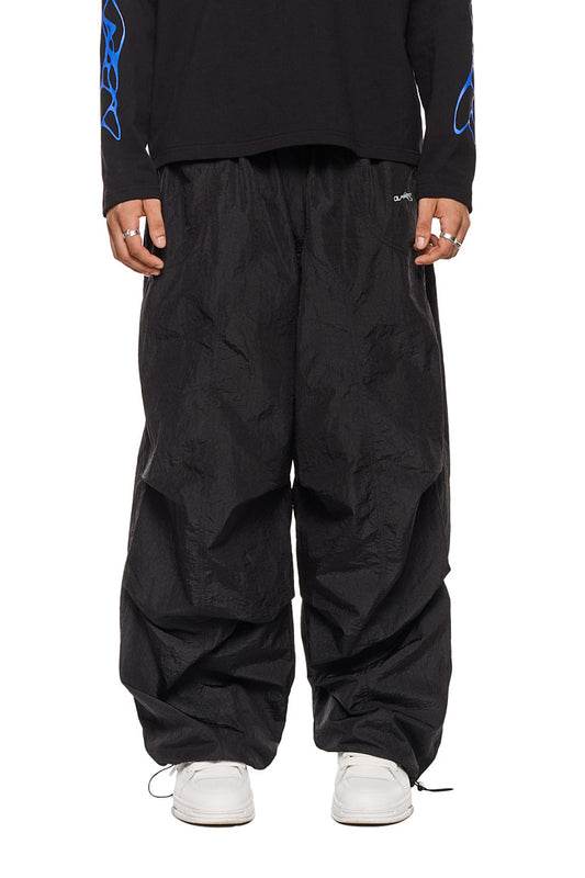 Person trägt schwarze Parachute Pants mit Seitentaschen, elastischem Kordelzug und gesticktem Synergy-Logo, aus strapazierfähigem Material von OLAKALA.