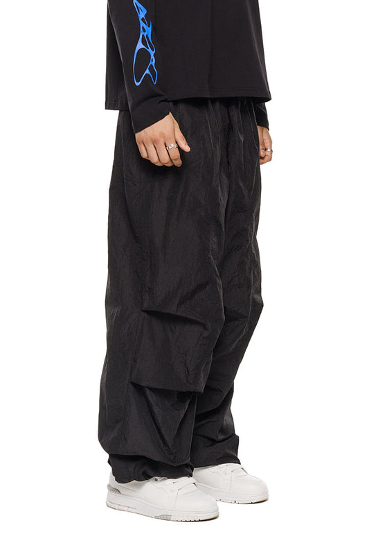 Person trägt schwarze Parachute Pants von OLAKALA mit elastischem Kordelzug, Eingriffstaschen und gesticktem Synergy-Logo, kombiniert mit weißen Schuhen.